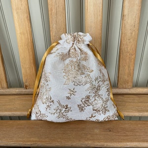 May include: A white drawstring bag with a gold floral pattern and gold satin ribbon ties.