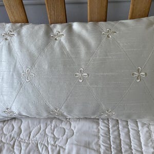 May include: A rectangular, off-white decorative pillow with a diamond pattern. The pillow features embroidered floral accents at the intersections of the diamond pattern. The pillow is resting on a white bedspread.