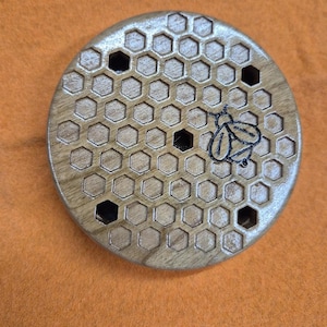 May include: A round wooden coaster with a honeycomb pattern and a small bee design. The coaster is made of light brown wood and has a natural finish.