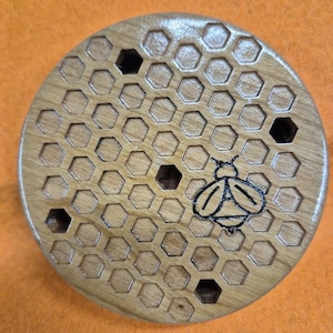 May include: A round wooden coaster with a honeycomb pattern and a black bee design. The coaster has several holes cut out of the honeycomb pattern.