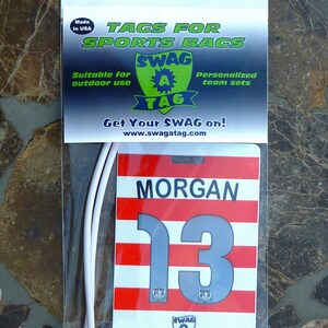 May include: White plastic bag with a red, white, and gray sports bag tag. The tag has the text "MORGAN" and the number "13" on it. The tag also has the text "SWAG A TAG" and the website "www.swagatag.com".