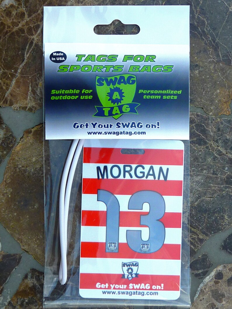 May include: White plastic bag with a red, white, and gray sports bag tag. The tag has the text "MORGAN" and the number "13" on it. The tag also has the text "SWAG A TAG" and the website "www.swagatag.com".