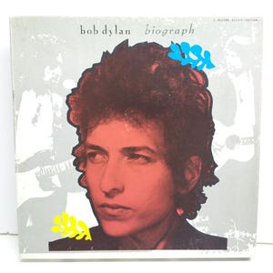 Puede incluir: Una portada de álbum de vinilo vintage con un retrato de cerca de Bob Dylan. El título del álbum "Biograph" está impreso en letras blancas sobre un fondo gris. La imagen está rodeada de formas abstractas de colores.