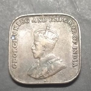 May include: A silver coin with a portrait of a crowned man, inscribed with "GEORGE V KING AND EMPEROR OF INDIA". The coin is square with rounded corners and shows signs of wear.