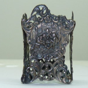 May include: Silver napkin ring with an ornate design featuring a scene of three people in a building. The ring is made of silver and has a delicate, intricate design.