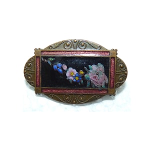 May include: Vintage oval brooch with a floral design. The brooch features a dark background with a painted floral arrangement in pink, blue, and yellow. The brooch is framed with a red border and a decorative bronze-colored metal.