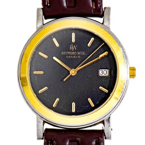 Könnte beinhalten: Eine zweifarbige Armbanduhr in Gold und Silber mit schwarzem Zifferblatt und braunem Lederband. Das Zifferblatt der Uhr hat goldene römische Ziffern als Stundenmarkierungen, goldene Zeiger und ein Datumsfenster bei 3 Uhr. Das Zifferblatt der Uhr trägt außerdem den Schriftzug "Raymond Weil Geneve" und "RW".