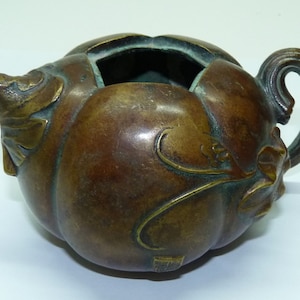 Puede incluir: Una tetera pequeña de bronce con forma de fruta, con asa y pico. La tetera tiene detalles intrincados y una pátina marrón oscura.