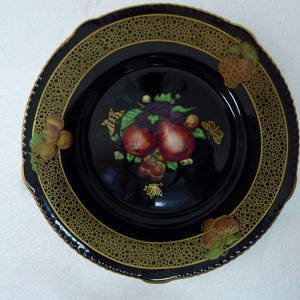 Könnte beinhalten: Schwarzer Keramikteller mit goldfarbenem Rand und einem Design aus Früchten und Schmetterlingen. Der Teller hat eine strukturierte Oberfläche.