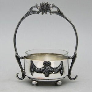 May include: A silver sugar bowl with a glass insert and a decorative handle in the shape of a flower. The bowl has a floral design on the front.