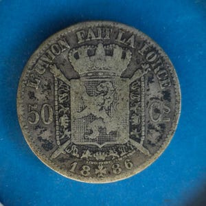 May include: A silver coin with the text "L'UNION FAIT LA FORCE" and the year "1886". The coin features a crowned crest with a lion and crossed swords. The number "50" and the letters "Cs" are also visible.