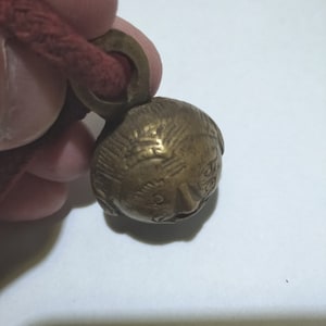 May include: A small, brass bell with a detailed face design. The bell is attached to a red cord.