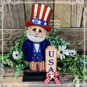 May include: A wooden figurine of a patriotic gnome wearing a red, white, and blue hat and a blue jacket with stars and stripes. He is holding a sign that says "USA" and is 11 inches tall.