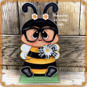 May include: A wooden cutout of a bee wearing glasses and a bow. The bee is painted yellow and black with white wings. The bee is holding a white daisy with a black and white checkered bow. The bee is 11 inches tall. "Ernie Made" is written on the bee's abdomen. "Measures 11 inches tall" is written on the wooden background.