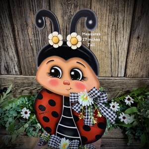 May include: A hand-painted wooden ladybug with black and white polka dots, red wings, and a black and white checkered bow. The ladybug has large eyes and a smiling face. It measures 17 inches tall. Measures 17 inches Tall.