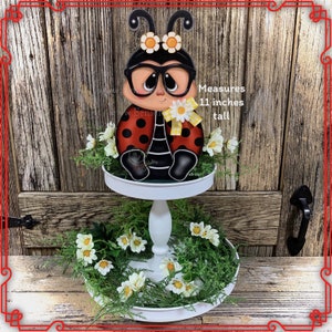 May include: A white two-tiered serving tray decorated with green leaves and white daisies. A ladybug figurine with black glasses and yellow accents sits on the top tier. The ladybug is 11 inches tall. Measures 11 inches tall.