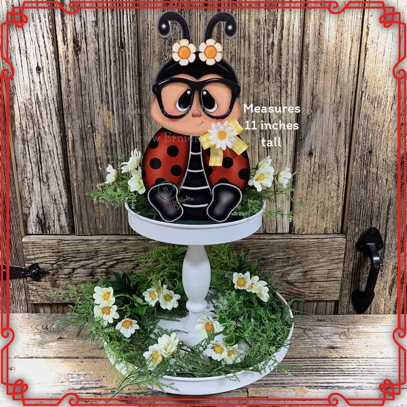 May include: A white two-tiered serving tray decorated with green leaves and white daisies. A ladybug figurine with black glasses and yellow accents sits on the top tier. The ladybug is 11 inches tall. Measures 11 inches tall.