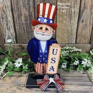 May include: A wooden figurine of Uncle Sam, painted in red, white, and blue, holding a sign that says "USA". The figurine is 17 inches tall and stands on a black base with a red, white, and blue bow.