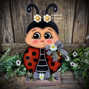 May include: A hand-painted wooden ladybug with black and red polka dots, black and white striped legs, and a black head with white eyes and yellow flowers. The ladybug is standing on a wooden base with a black and white checkered ribbon and white flowers. The ladybug measures 17 inches tall. "Ernie Mae" is written on the ladybug's body.