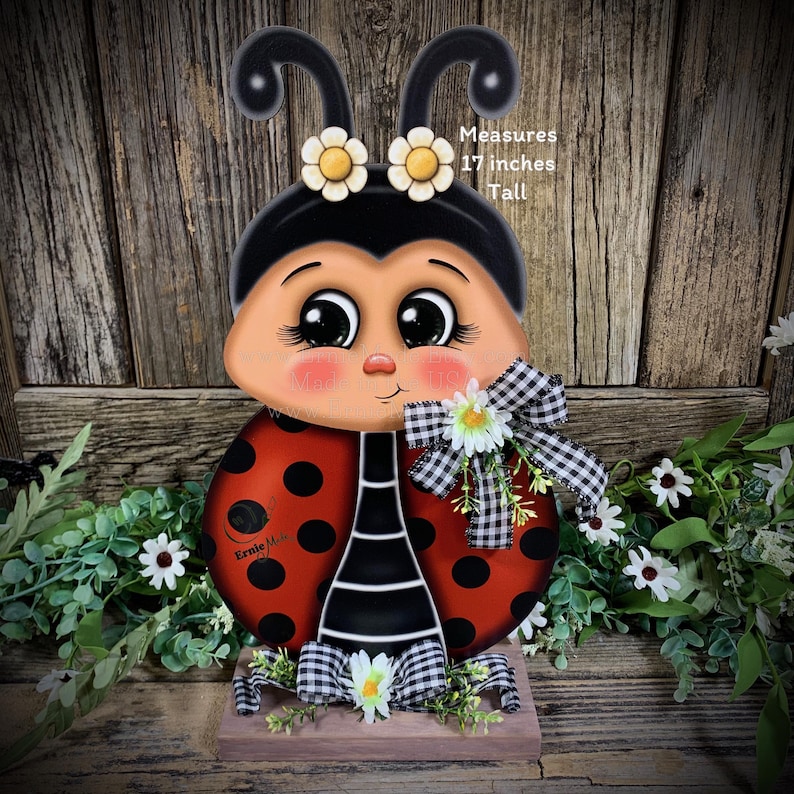 May include: A hand-painted wooden ladybug with black and red polka dots, black and white striped legs, and a black head with white eyes and yellow flowers. The ladybug is standing on a wooden base with a black and white checkered ribbon and white flowers. The ladybug measures 17 inches tall. "Ernie Mae" is written on the ladybug's body.