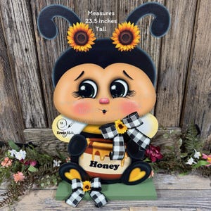 May include: A whimsical, wooden bee decoration, 23.5 inches tall, with a black and yellow body, large eyes, and a sad expression. It features sunflower accents, a honey pot, and a black and white checkered bow. The base is green.