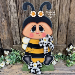 May include: A wooden bee decoration, 23.5 inches tall, with a black and yellow striped body, large eyes, and a sweet expression. It has black and white checkered bows and daisy accents. The text "Ernie Made" is visible.