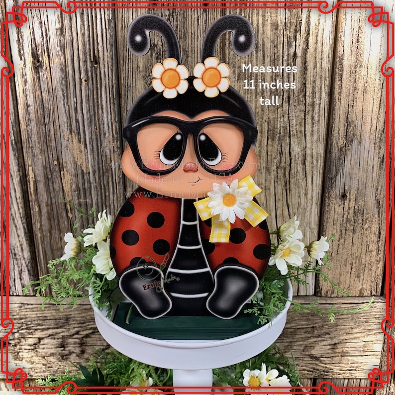 May include: A ladybug figurine with black and white striped legs, red wings with black dots, and large black glasses. The ladybug is wearing a yellow and white bow and has yellow flowers on its head. The figurine is 11 inches tall and is sitting on a white tiered stand with green foliage and white flowers.