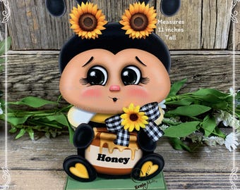 May include: A wooden cutout of a bee holding a jar of honey. The bee is painted with black and yellow stripes, has large brown eyes, and is wearing a black and white checkered bow. The jar of honey is painted brown and has the word "Honey" written on it. The bee is 11 inches tall.