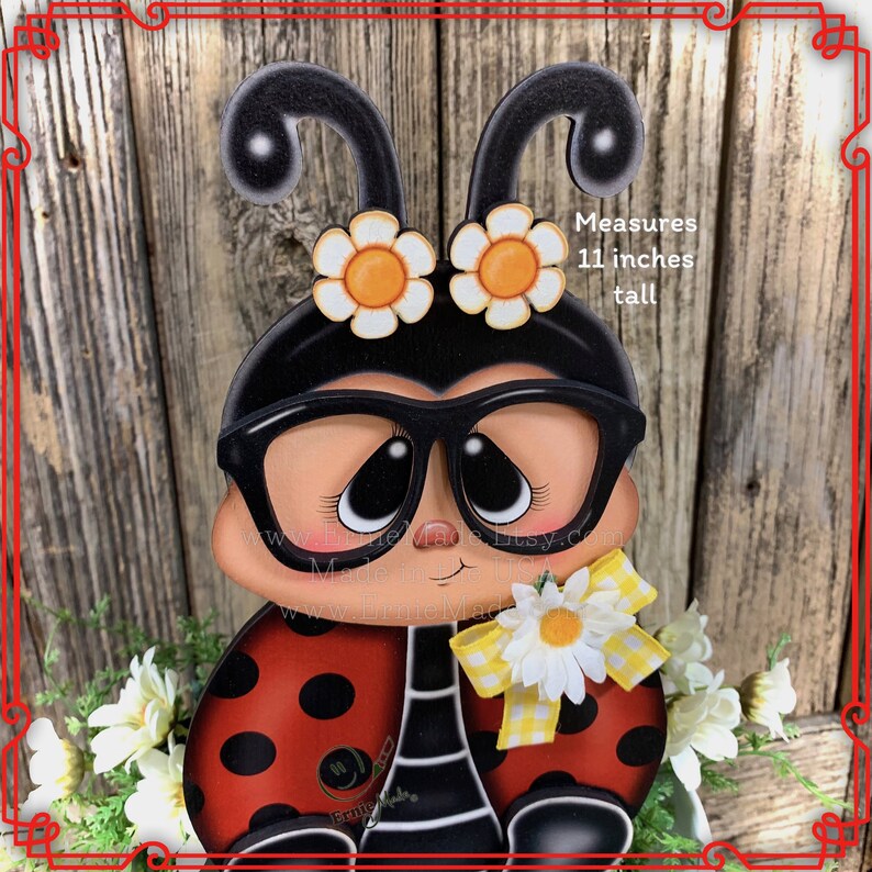 May include: A wooden ladybug figurine with black glasses, red and black polka dots, and a white daisy with a yellow ribbon. It measures 11 inches tall. "Measures 11 inches tall" is written on the wooden background.