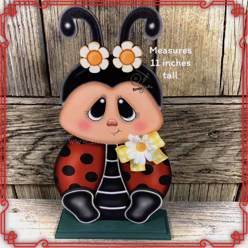 May include: A hand-painted wooden ladybug figurine with black and white polka dots, a yellow bow, and a white daisy. The ladybug is sitting on a green base and measures 11 inches tall. "Measures 11 inches tall" is written in black text on the right side of the image.