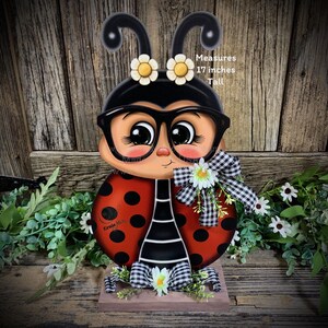 May include: A wooden ladybug figurine with black and white stripes, red body with black dots, and large black-rimmed glasses. The ladybug is wearing a black and white checkered bow and has white flowers on its head. The figurine is 17 inches tall.
