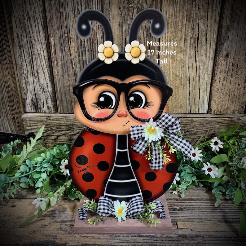 May include: A wooden ladybug figurine with black and white stripes, red body with black dots, and large black-rimmed glasses. The ladybug is wearing a black and white checkered bow and has white flowers on its head. The figurine is 17 inches tall.