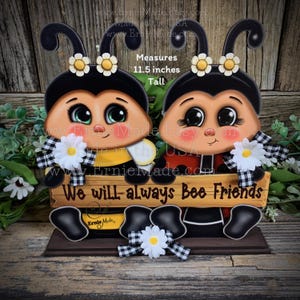 May include: A wooden bee-themed decoration featuring two cartoon bees with black and white accents, holding a sign that reads "We will always Bee Friends." The decoration measures 11.5 inches tall.