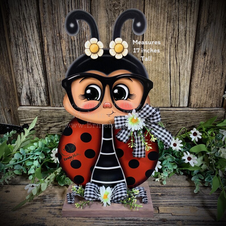 May include: A hand-painted wooden ladybug with black and white polka dots, wearing glasses and a black and white checkered bow. The ladybug is 17 inches tall and has a red body with black spots. The ladybug is standing on a wooden base with green foliage and white flowers.