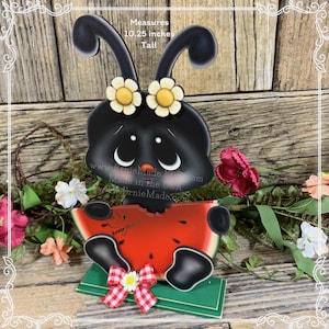 K&ouml;nnte beinhalten: Schwarze und wei&szlig;e Marienk&auml;fer-Figur, die ein St&uuml;ck Wassermelone h&auml;lt. Der Marienk&auml;fer tr&auml;gt gelbe Blumen und eine rote Schleife. Die Figur ist 26 cm hoch. "Ernie Made" steht auf dem Wassermelonenst&uuml;ck.