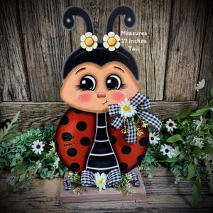 May include: A hand-painted wooden ladybug figurine with black and white stripes, red body with black dots, and a black head with large eyes. The ladybug is wearing a white and black checkered bow and has white flowers on its head. The figurine is 17 inches tall and stands on a wooden base.