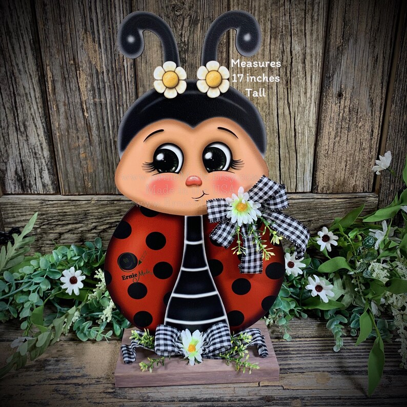 May include: A hand-painted wooden ladybug figurine with black and white stripes, red body with black dots, and a black head with large eyes. The ladybug is wearing a white and black checkered bow and has white flowers on its head. The figurine is 17 inches tall and stands on a wooden base.