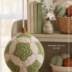 May include: A handmade crochet pendant light with a green and cream geometric pattern. The light hangs from a rope and is suspended above a shelf with books, yarn, and decorative items. The text "Handmade in the USA Est. 2024" is visible.