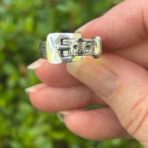 May include: A silver ring with a buckle design. The ring features a square buckle with detailed patterns and a small, rounded clasp. The ring is held against a blurred green background.