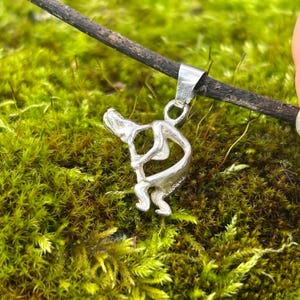 May include: A silver pendant shaped like a person playing a musical instrument. The pendant is hanging from a branch over a bed of green moss.