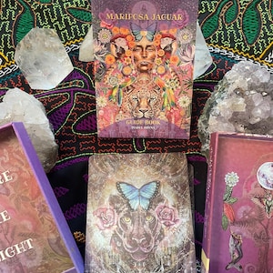 May include: A Mariposa Jaguar guide book and tarot card deck with a purple box. The book cover features a woman's face, a jaguar, and a butterfly. The cards show a jaguar, butterfly, and roses. The background is a colorful, patterned fabric.