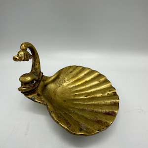May include: A vintage brass swan-shaped dish with a shell-like bowl. The swan's neck curves upward, and its head forms a small handle. The dish has a weathered, antique appearance, with visible patina and detailed textures.