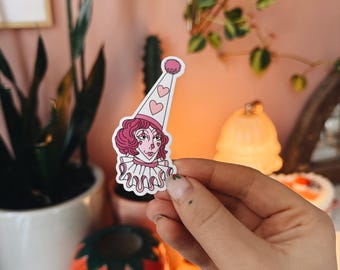 May include: A pink and white sticker of a clown with a pink and white hat with three pink hearts on it. The clown has pink hair and a pink collar.