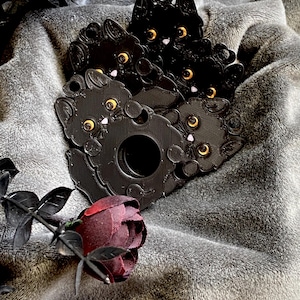 May include: A collection of black cat-shaped charms featuring textured surfaces, prominent orange eyes, and small pink noses. Each charm includes a circular cutout and a small loop at the top for attachment, arranged in a cluster with a dark red rose.