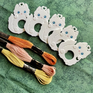 May include: Five white cat-shaped plastic thread bobbins with blue eyes and pink noses, arranged in a row. Below the bobbins are three skeins of embroidery floss in orange, light pink, and yellow, each secured with a black paper label.