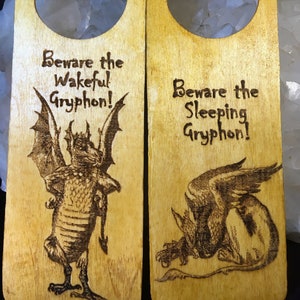 May include: Two wooden door hangers with a light brown finish. Each has a teardrop-shaped hole at the top. One features a detailed illustration of a gryphon with the text "Beware the Wakeful Gryphon!". The other says "Beware the Sleeping Gryphon!"