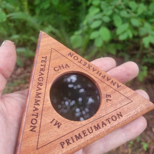 Pode incluir: Um pingente triangular de madeira com uma janela circular no centro. O pingente é gravado com as palavras "Tetragrammaton", "CHA", "Anaphaxeton", "EL" e "Primeumaton".