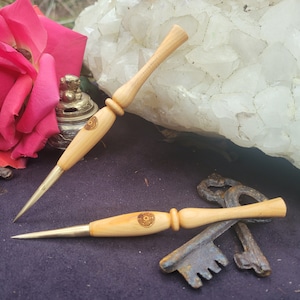 May include: Two wooden tools with a brass tip and a circular design burned into the wood. The tools are lying on a dark brown surface with a rusty key in the background.