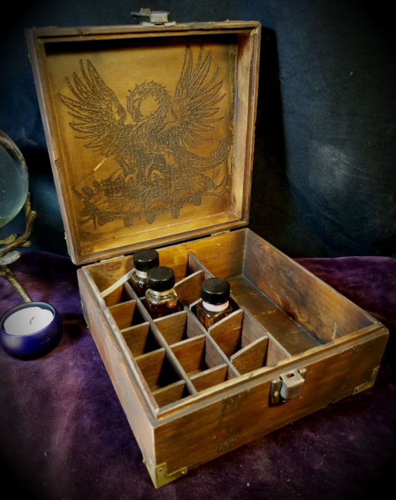 May include: A wooden box with a hinged lid, featuring an engraved image of a griffin on the inside of the lid. The box has multiple compartments inside, and four small glass bottles are visible in the compartments.