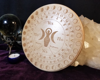 May include: A circular wooden divination board, engraved with letters, numbers, and moon phases. The central design features a female figure between two crescent moons. The board includes the words "Yes", "No", "Maybe", and "Rephrase".